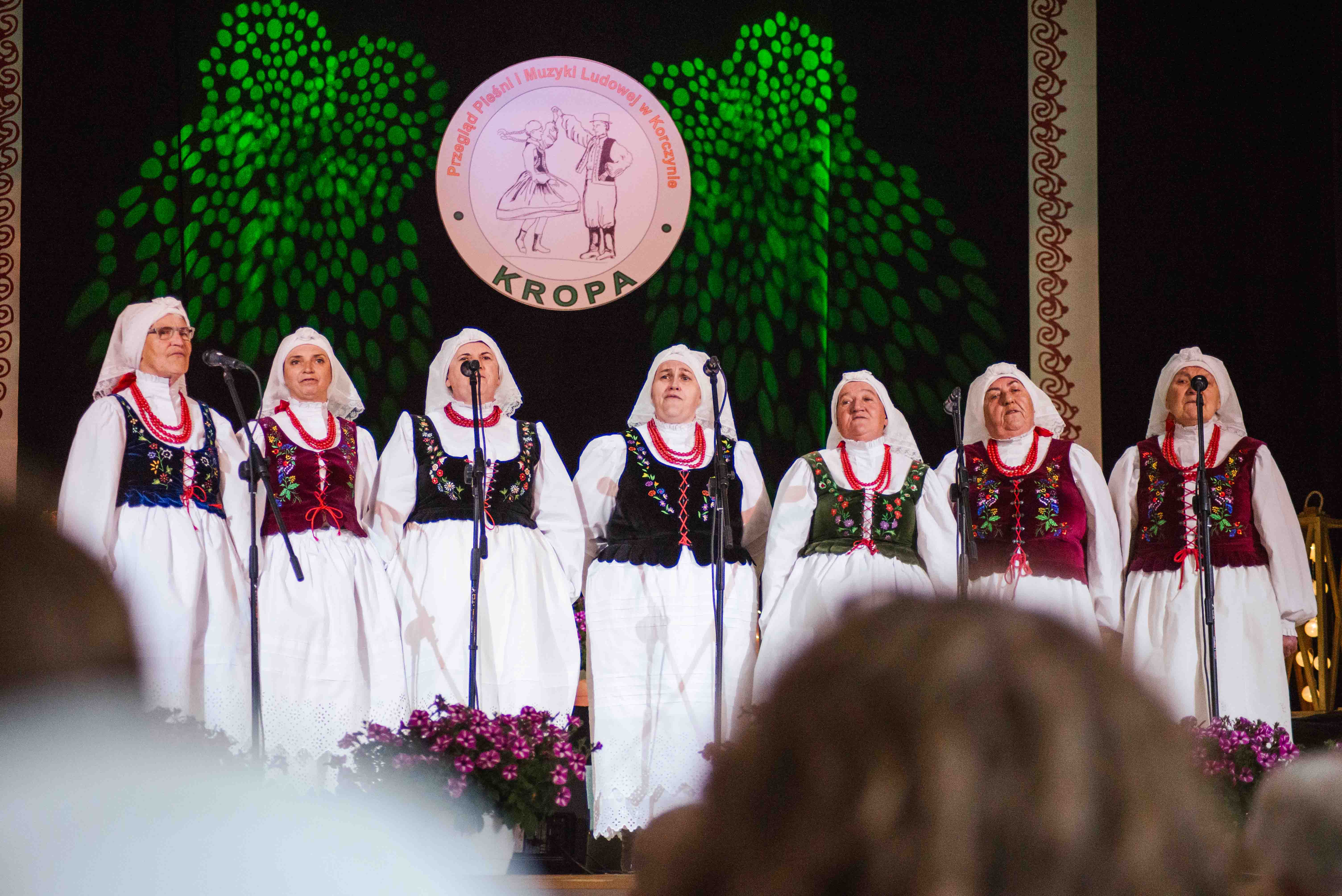 Rząd siedmiu kobiet w strojach ludowych śpiewających na scenie.
